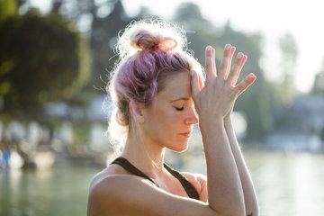 Die Aufnahme zeigt eine Person am Wasser, die eine ruhige, konzentrierte Haltung mit erhobenen Händen einnimmt. Im Hintergrund sind Bäume und weiches Sonnenlicht zu sehen, die eine natürliche, friedliche Stimmung erzeugen.